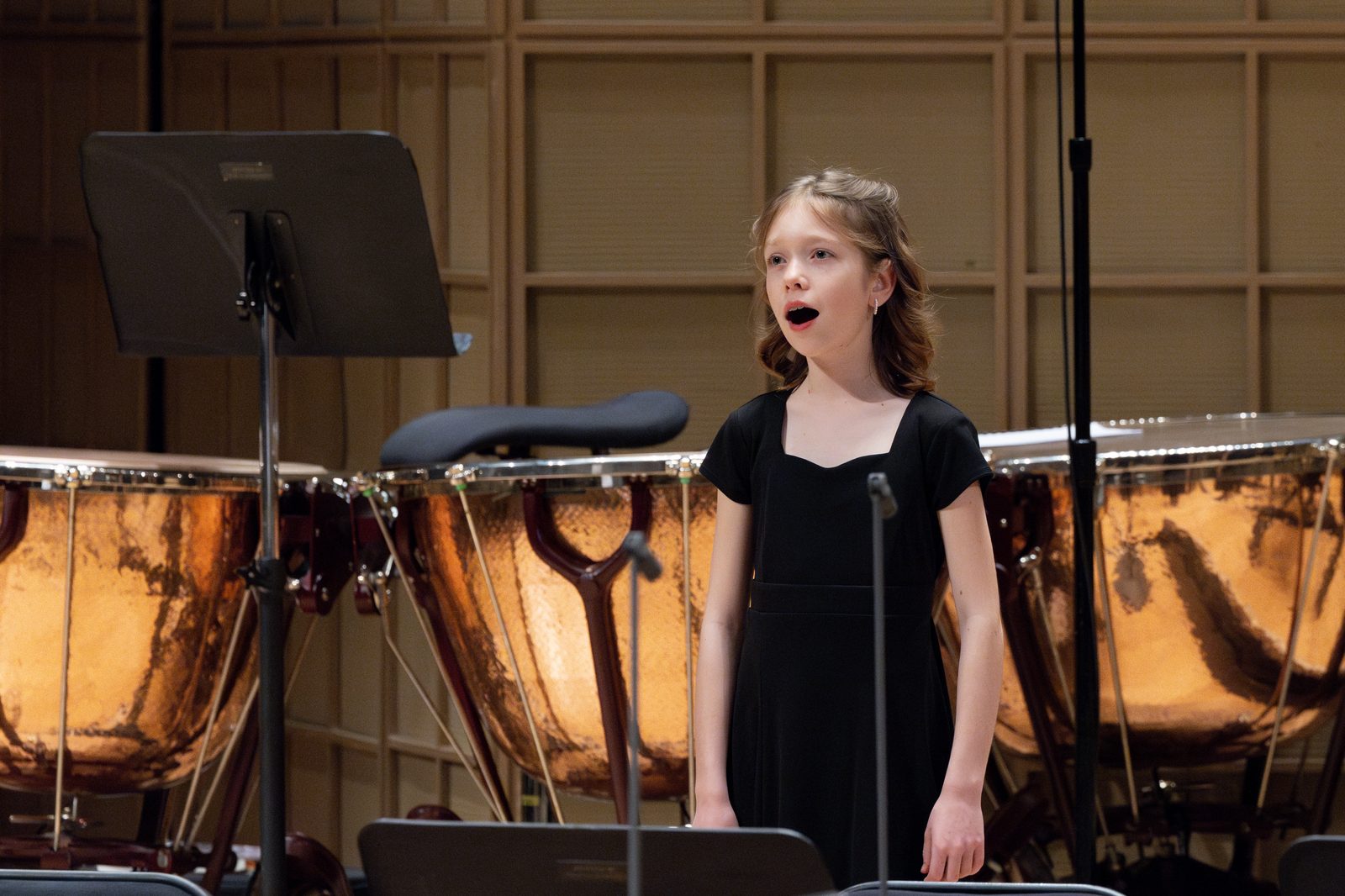 A young chorister in a black concert dress singing on stage, with the orchestra&rsquo;s copper timpani behind her