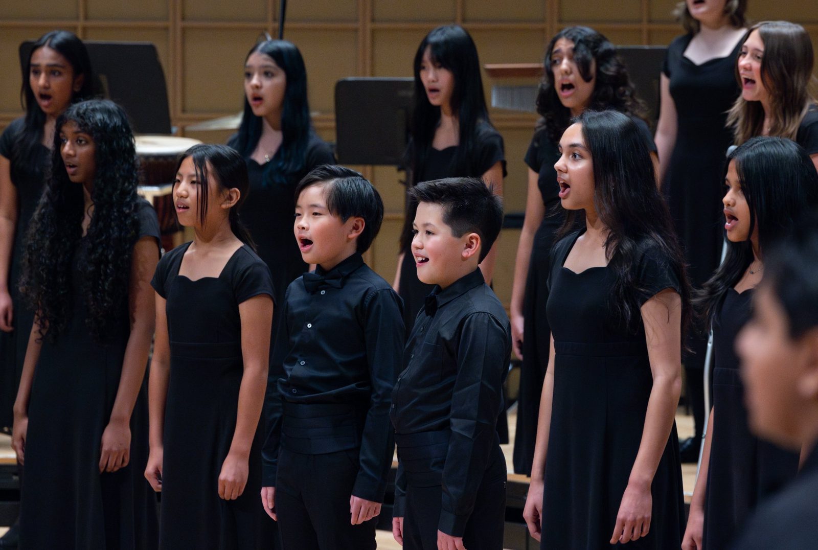 Choristers in black concert attire singing together on stage, with two younger boys in bow ties at the front and older singers on the risers behind them